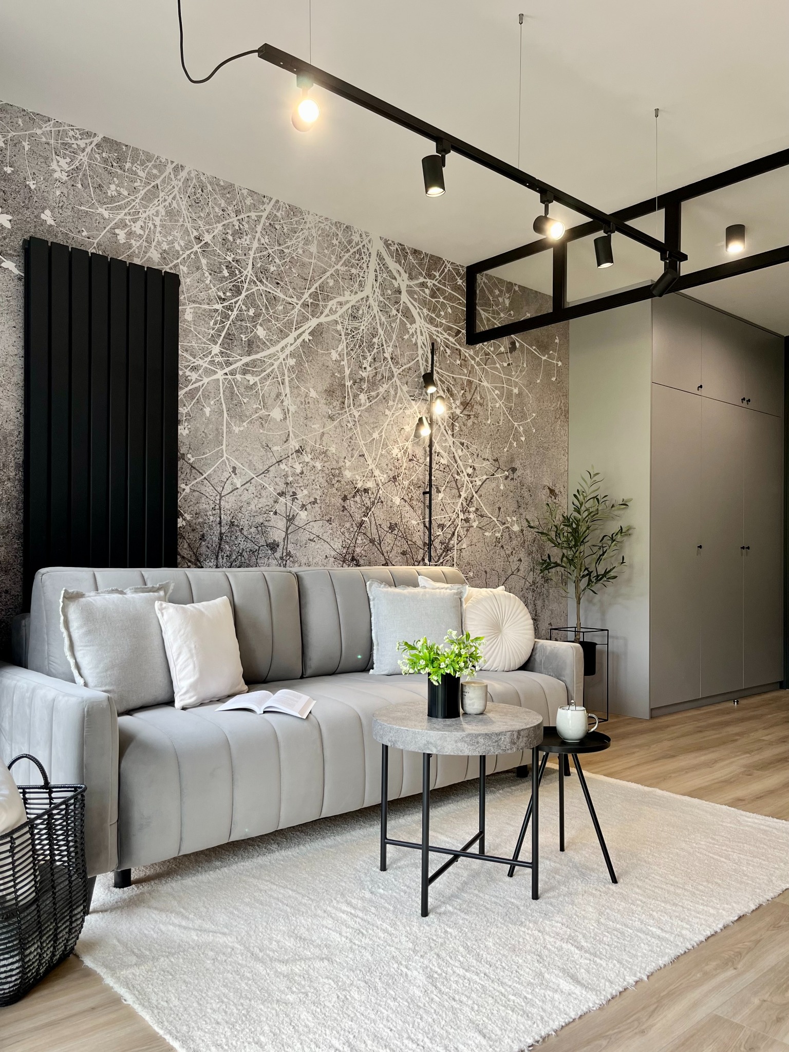 Stylowy salon w Krakowie z szarym, welurowym narożnikiem, okrągłymi stolikami kawowymi z marmurowym blatem, wzorzystą tapetą imitującą gałęzie drzew i czarnym grzejnikiem panelowym.