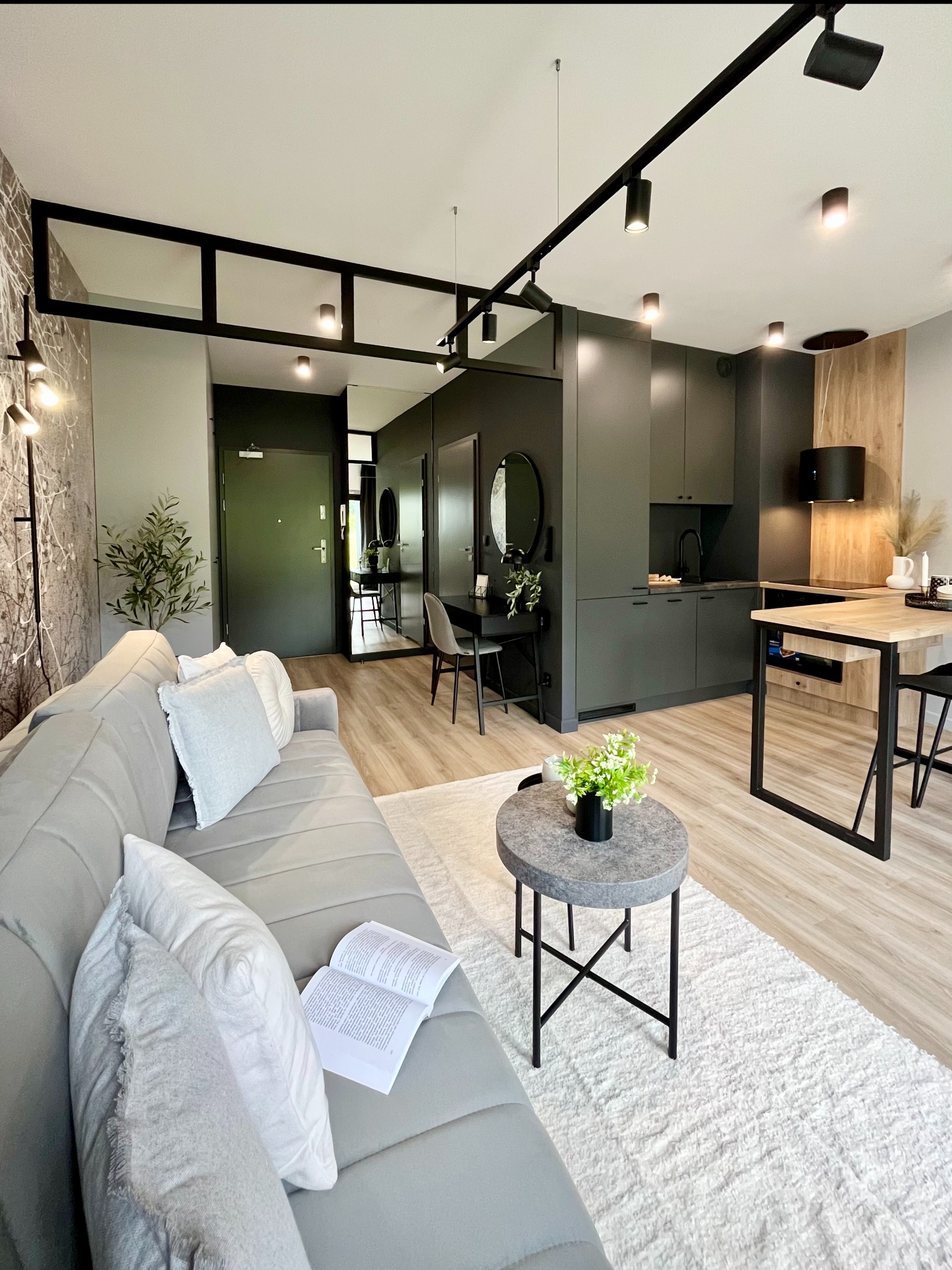 Nowoczesne studio: szary narożnik z poduszkami i otwartą książką, okrągły stolik z kwiatami, kuchnia w odcieniach szarości z drewnianym blatem, czarne oświetlenie szynowe.