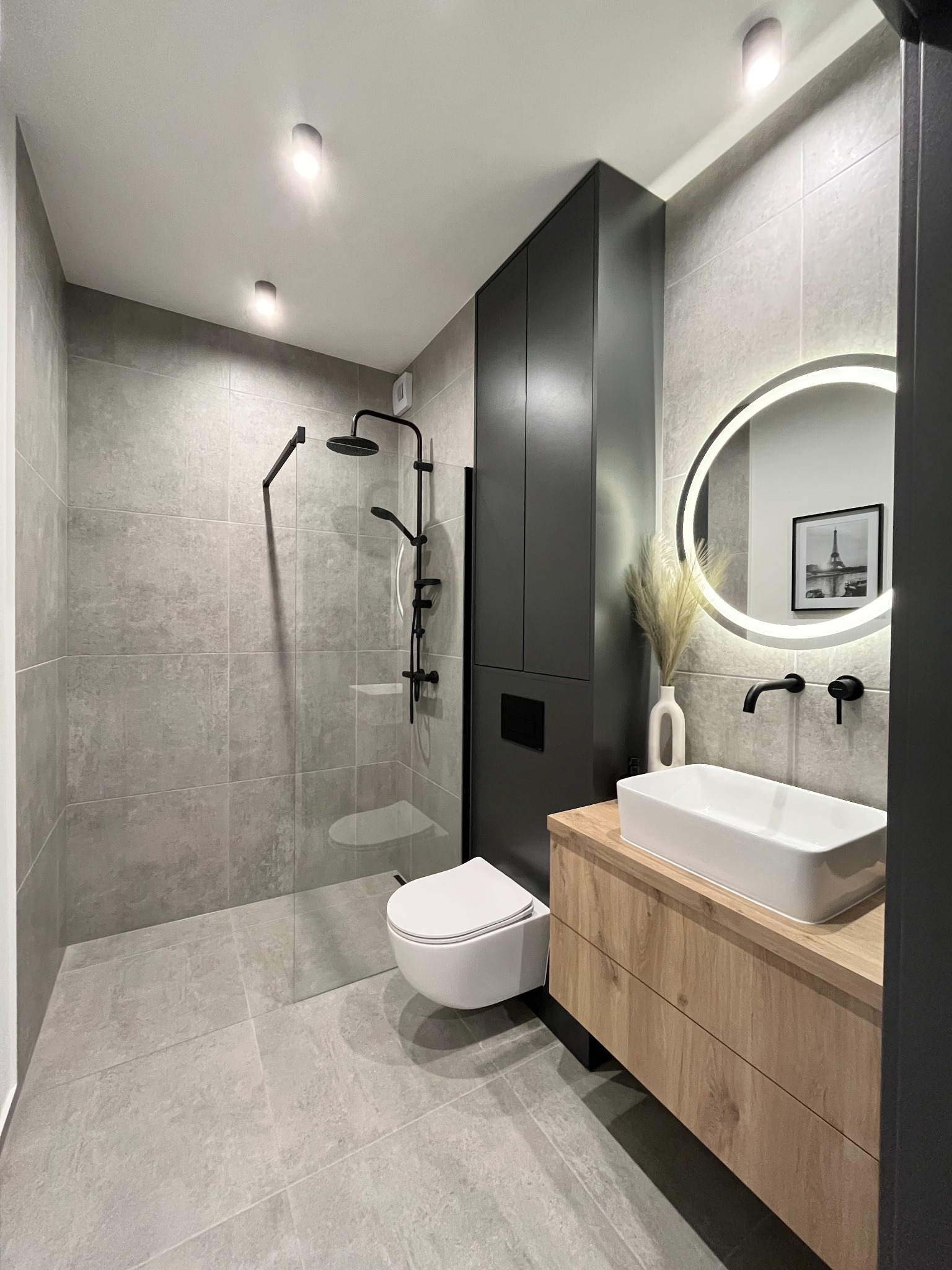 Nowoczesna łazienka w szarościach z akcentami czerni i drewna: kabina prysznicowa typu walk-in, podświetlane okrągłe lustro nad umywalką nablatową, szafka z drewnianym frontem i czarny słupek.