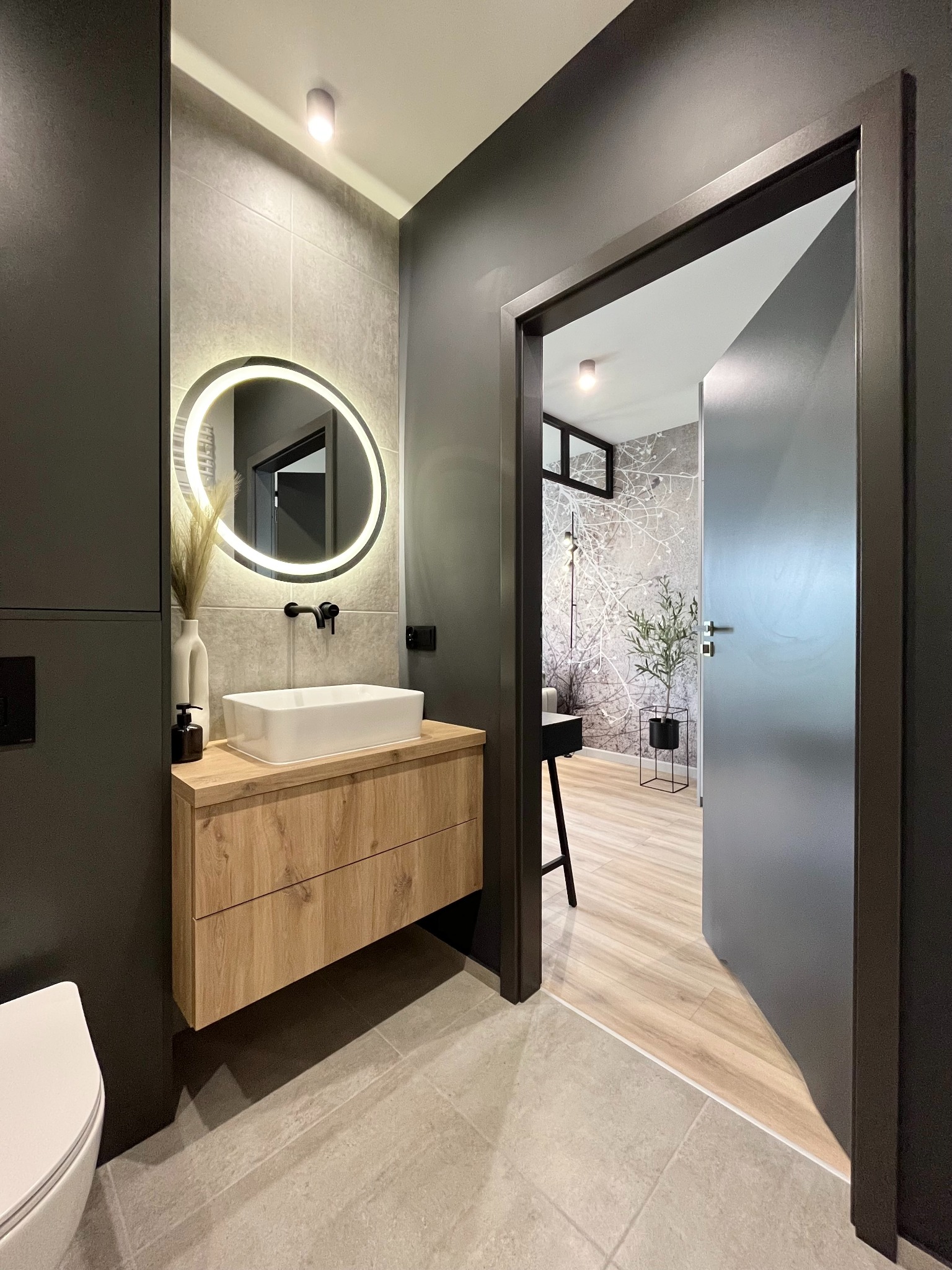 Nowoczesna łazienka w odcieniach szarości z drewnianą szafką pod umywalką, okrągłym lustrem z podświetleniem LED i widokiem na pokój z tapetą w motywy roślinne.