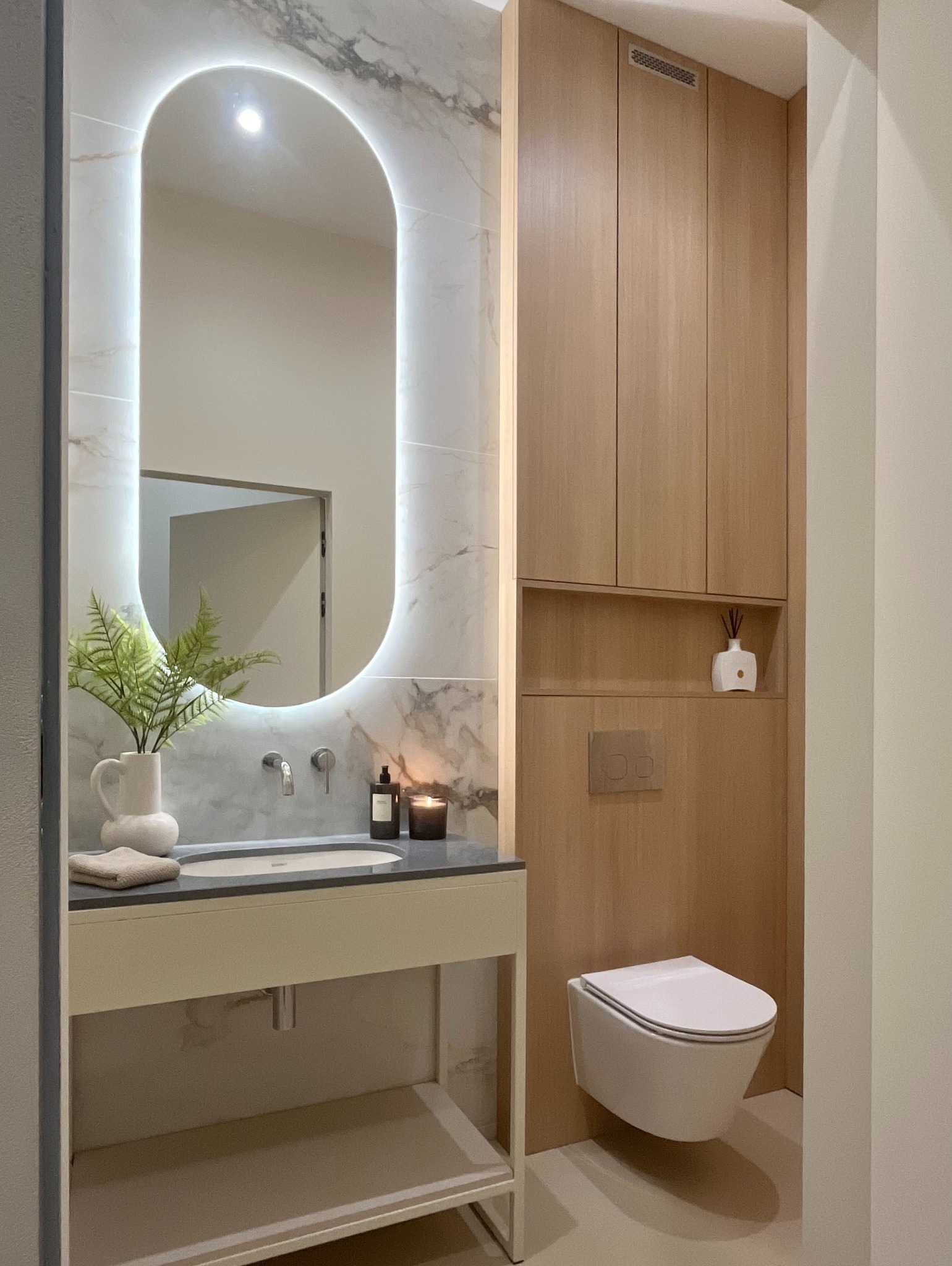 Nowoczesna łazienka z podświetlanym lustrem o zaokrąglonym kształcie, marmurową ścianą, drewnianymi szafkami, wiszącą toaletą i blatem z umywalką, uzupełniona o roślinę w wazonie i świecę.