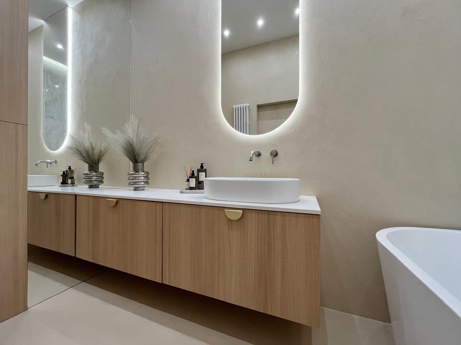 Nowoczesna łazienka z drewnianą szafką pod umywalką, owalnym lustrem z podświetleniem LED i dekoracyjnymi wazonami z trawą pampasową. Minimalistyczny design, ciepła kolorystyka.