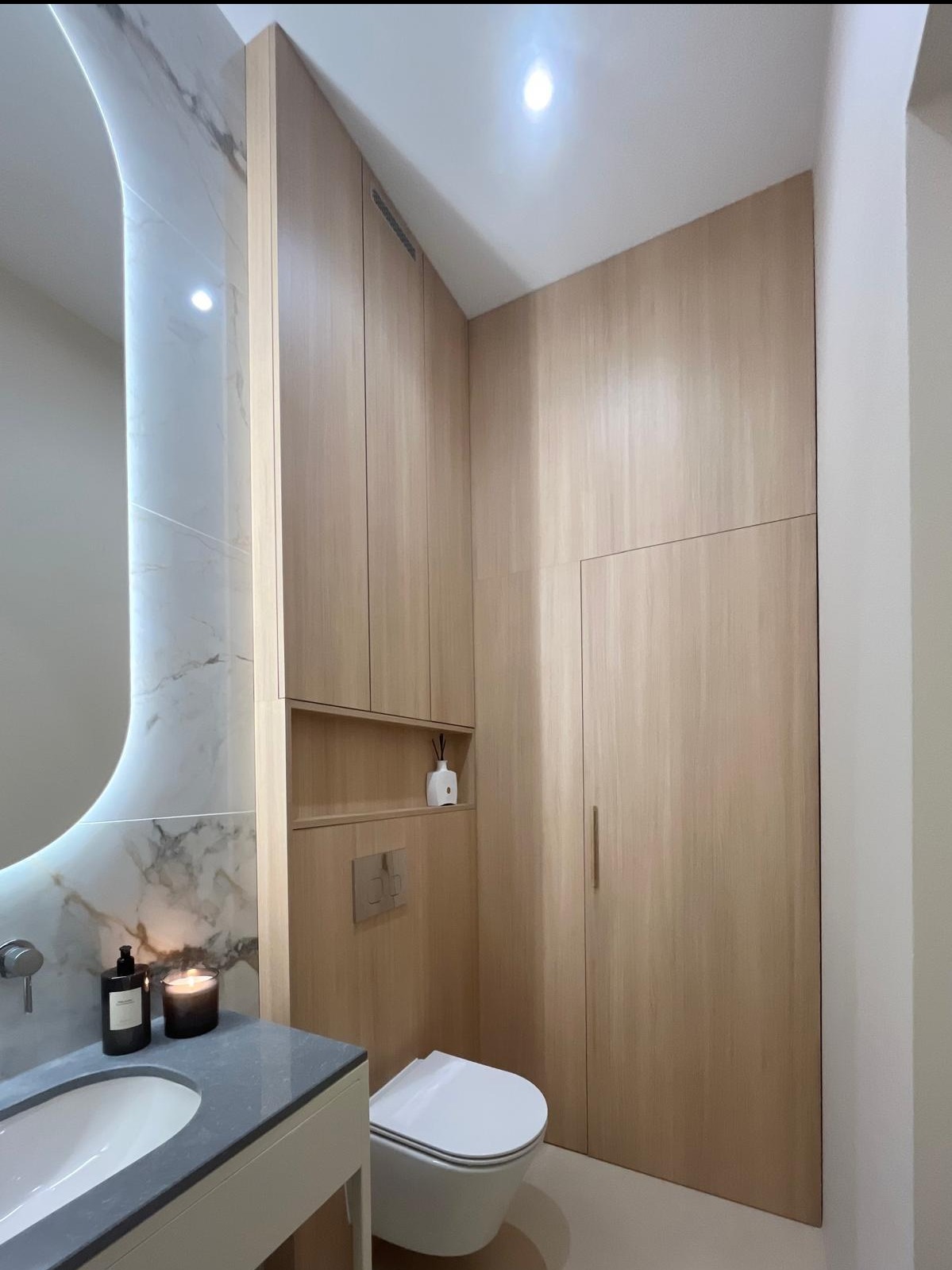 Nowoczesna łazienka z zabudową meblową z jasnego drewna i marmurową ścianą z podświetlanym lustrem. Minimalistyczny design, ciepłe oświetlenie, wisząca toaleta.