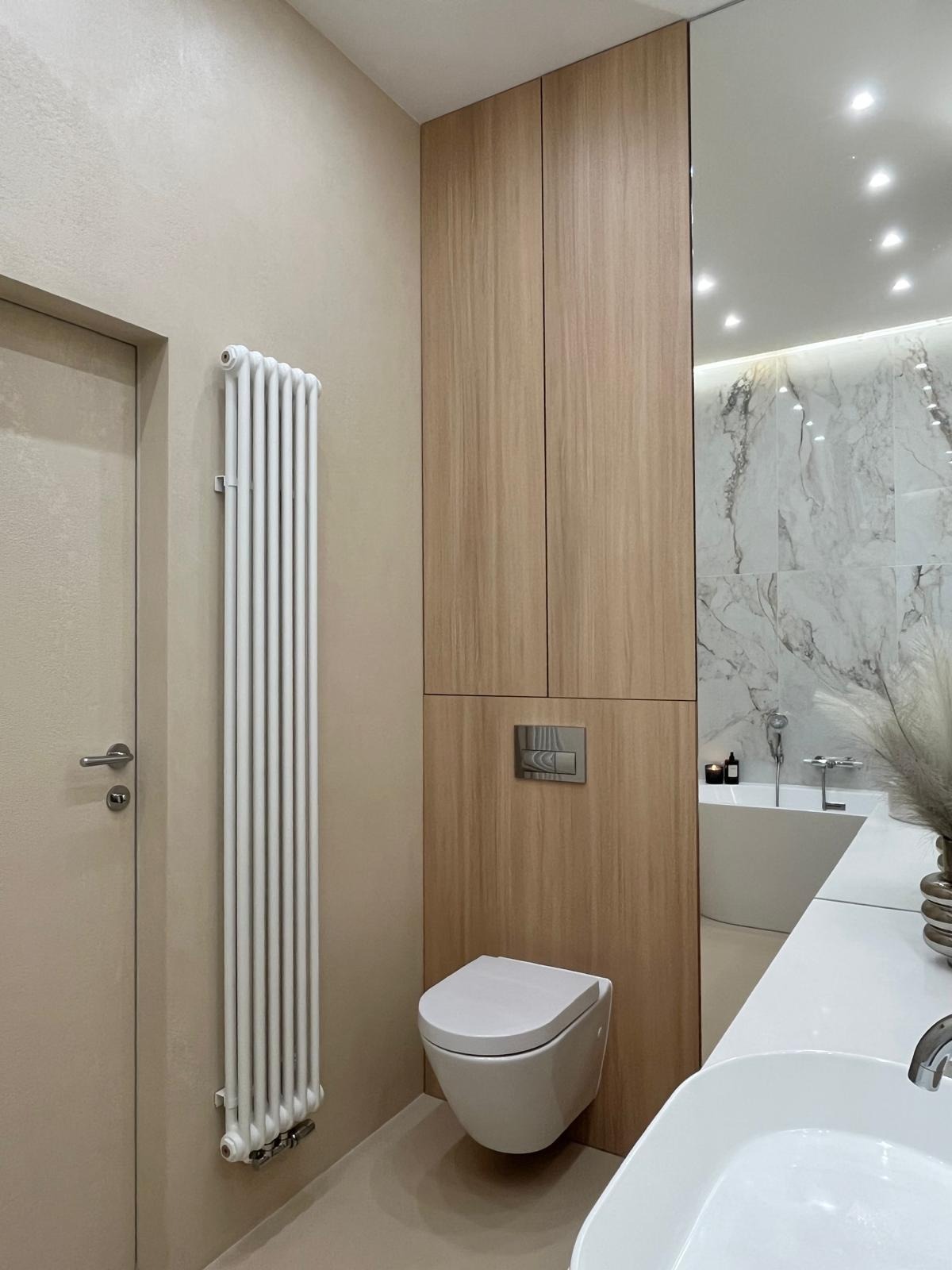 Nowoczesna łazienka z beżowymi ścianami, grzejnikiem żeberkowym, podwieszaną toaletą i drewnianą szafką. W lustrze odbija się wanna i marmurowe płytki.