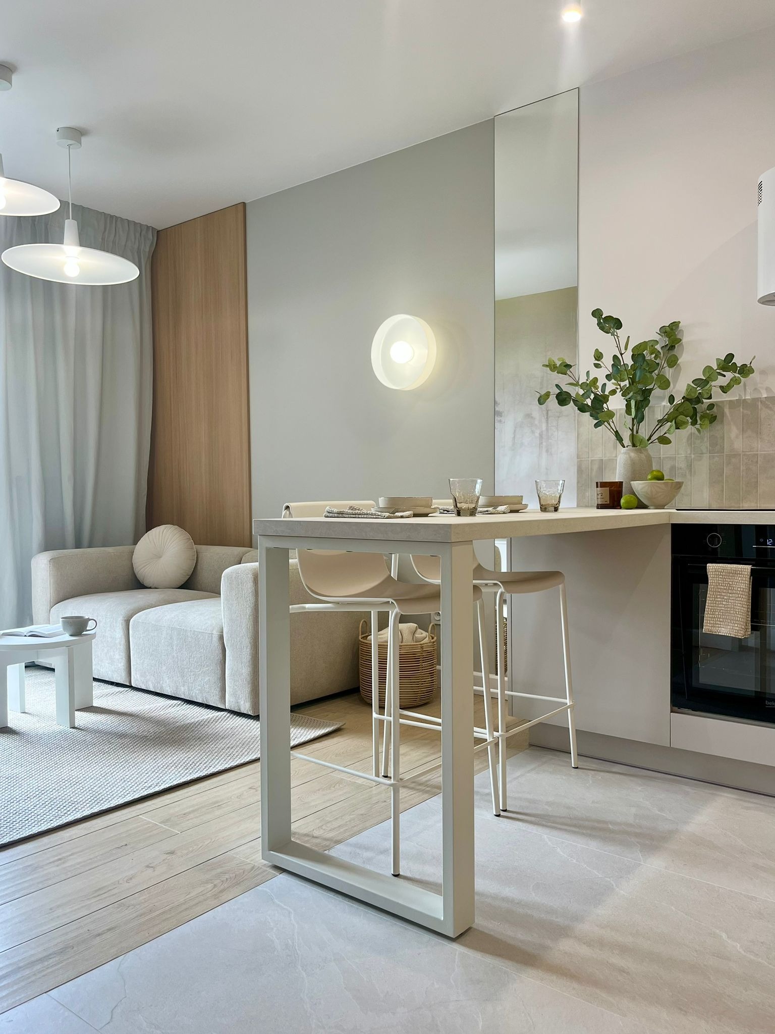Jasna aranżacja wnętrza: salon z aneksem kuchennym. Minimalistyczny design, beżowa sofa, biały stół z krzesłami barowymi i subtelne dodatki roślinne.