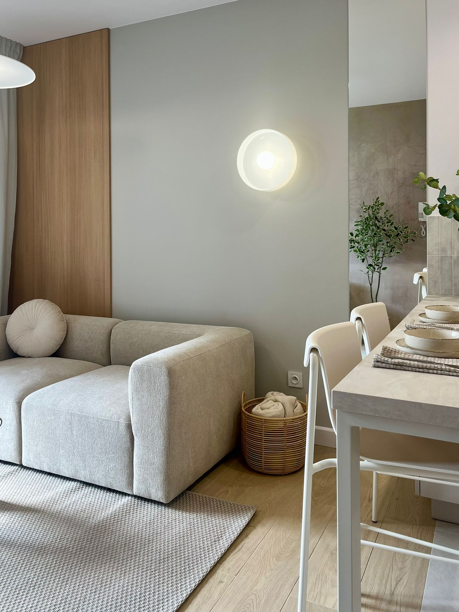 Jasne wnętrze: beżowa sofa z okrągłą poduszką, drewniana boazeria, okrągła lampa na ścianie, stół z nakryciem i krzesła. Neutralna kolorystyka, minimalistyczny wystrój.
