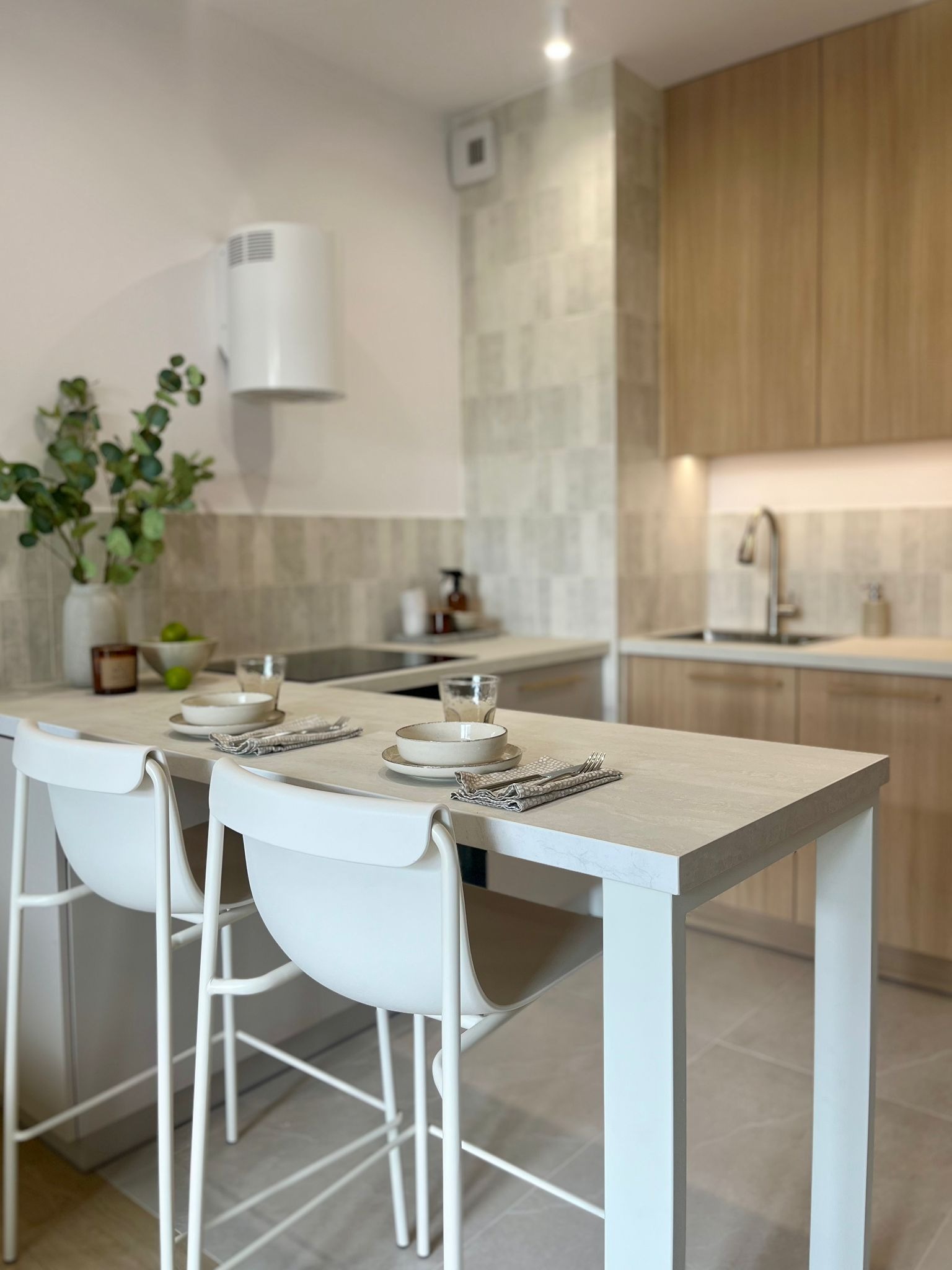 Jasna kuchnia z wyspą i białymi hokerami. Na blacie zastawa, w tle płytki imitujące kamień i drewniane szafki. Minimalistyczny okap cylindryczny.