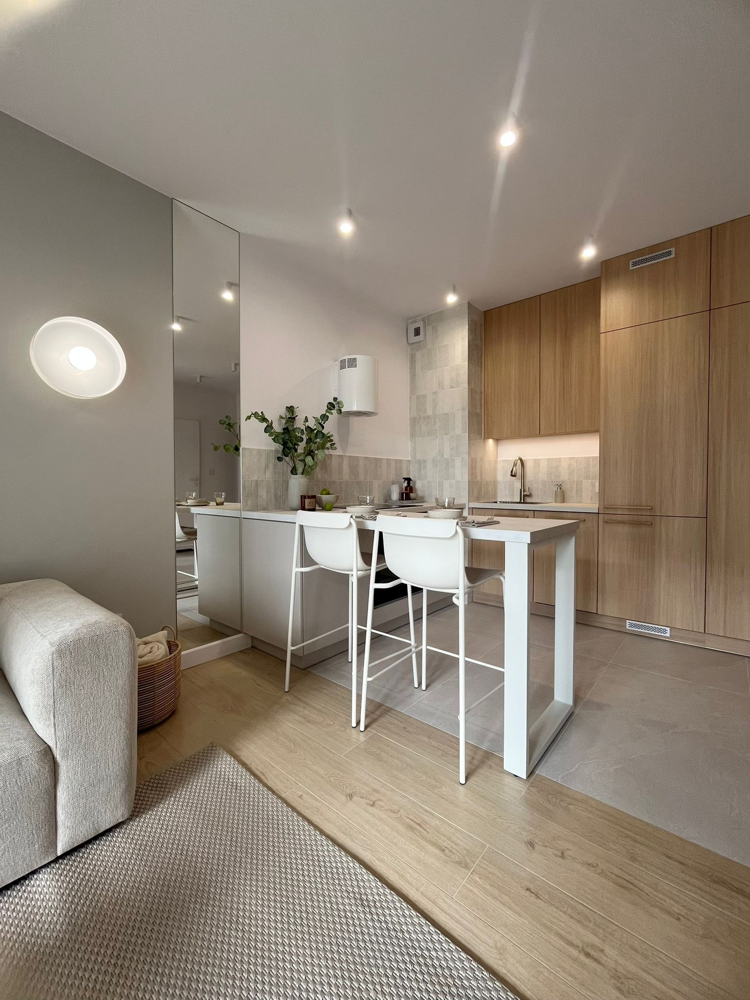 Jasna kuchnia z drewnianymi szafkami, białymi krzesłami barowymi i lustrem. Minimalistyczny design z roślinami w wazonie. Widoczny fragment sofy i dywanu.