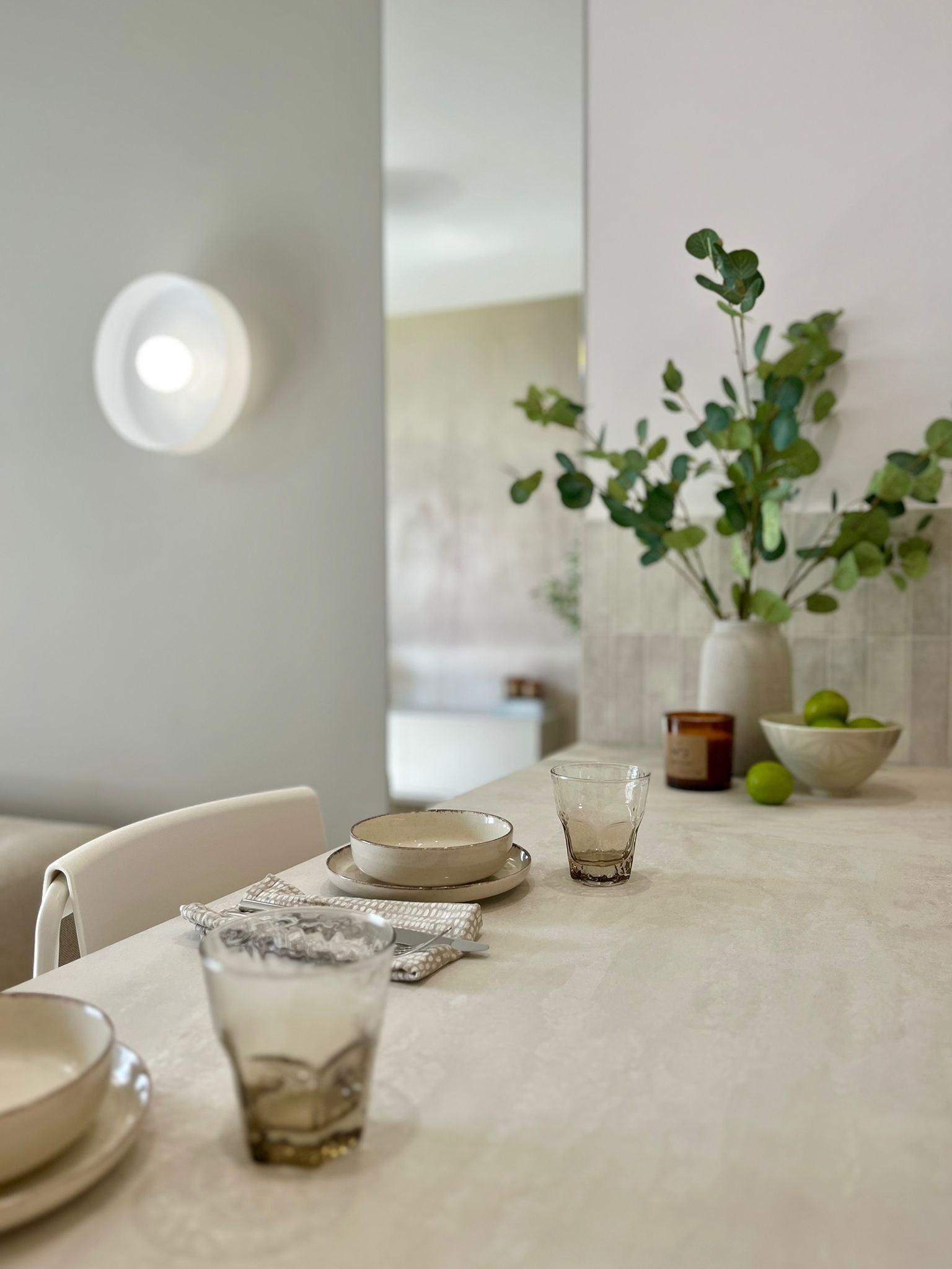 Minimalistyczna aranżacja stołu w jasnej tonacji: ceramiczne naczynia, szklanki, świeca i wazon z eukaliptusem na tle ściany z oświetleniem punktowym.