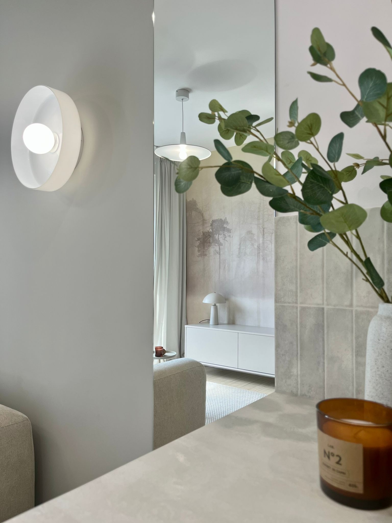 Minimalistyczny salon z dekoracyjnym oświetleniem ściennym,  rośliną w wazonie i świecą. W tle widoczne lustro odbijające wnętrze pokoju.