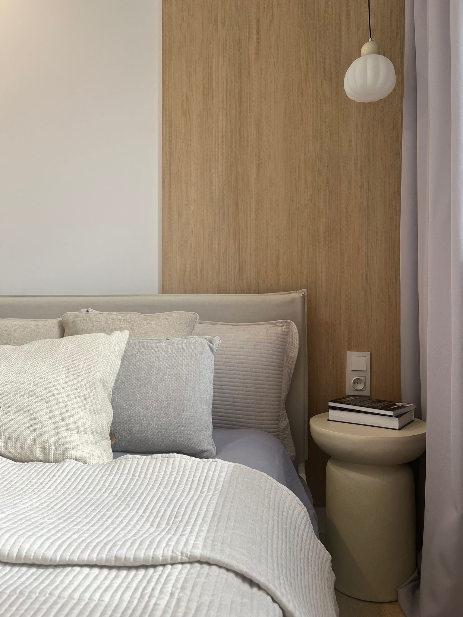 Neutralna sypialnia: beżowe drewno, szara pościel, lampka nocna i książki na okrągłym stoliku. Minimalistyczny design wnętrza z delikatnym oświetleniem.