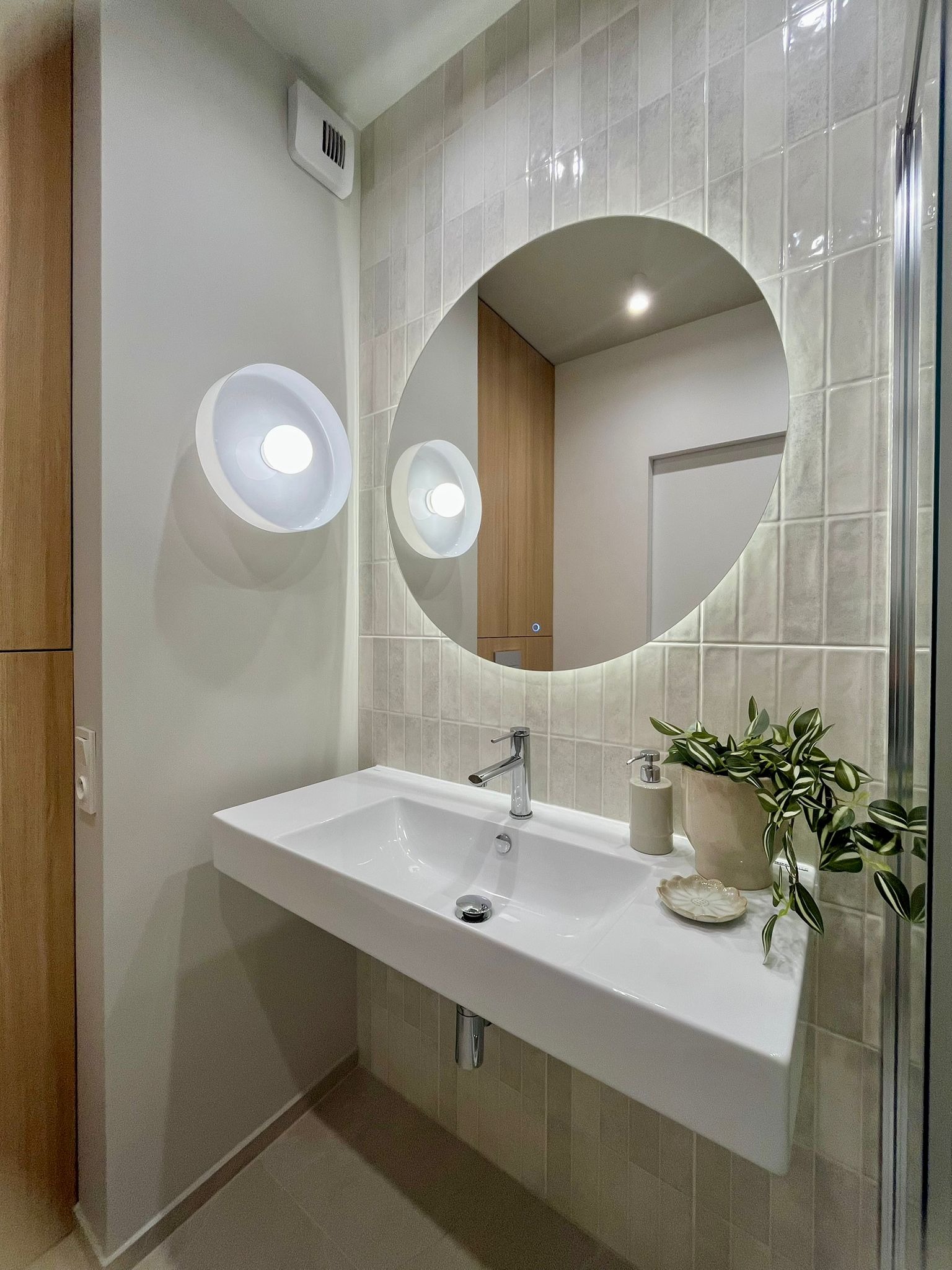 Nowoczesna łazienka z umywalką nablatową, okrągłym lustrem z podświetleniem LED i płytkami w odcieniach beżu. Roślina doniczkowa dodaje świeżości.