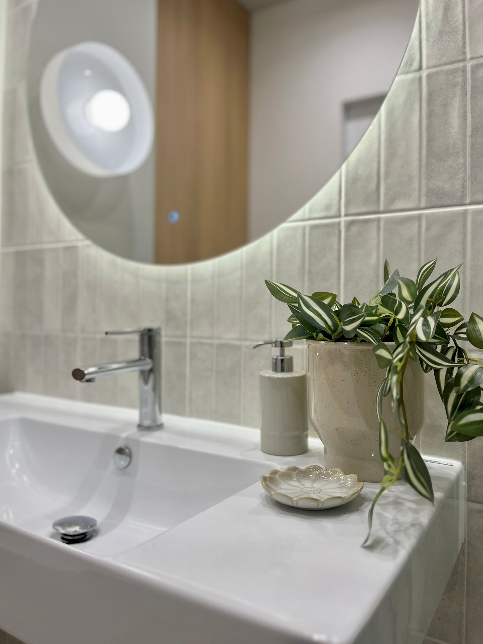 Nowoczesna łazienka: biała umywalka z chromowaną baterią, okrągłe lustro z podświetleniem, ceramiczna doniczka z zieloną rośliną i mydelniczka w kształcie kwiatka.