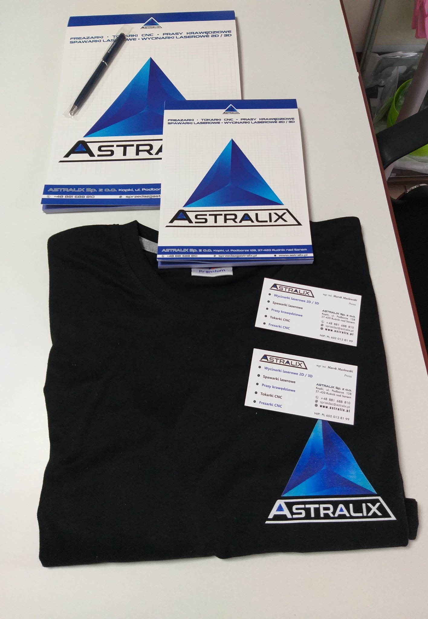 Materiały reklamowe firmy Astralix: notatniki, wizytówki i czarny t-shirt z logo, prezentujące usługi wycinania laserowego, spawania laserowego, pras krawędziowych, tokarek CNC i frezarek CNC.