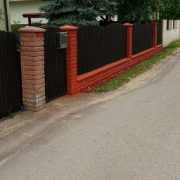 Tom-Bud Uslugi remontowo budowlane - Ciemnobrązowe drewniane ogrodzenie z ceglanymi słupkami pomalowanymi na czerwono, z metalową skrzynką na listy, na tle zielonego drzewa i domu z ciemną dachówką.