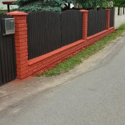 Tom-Bud Uslugi remontowo budowlane - Odnowiony, pomalowany na czerwono ceglany murek i ciemne drewniane panele ogrodzenia przy ulicy, z widoczną skrzynką pocztową i zielenią w tle.
