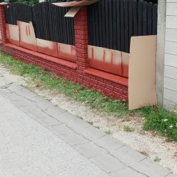 Tom-Bud Uslugi remontowo budowlane - Częściowo pomalowane ogrodzenie z czarnymi deskami i czerwonymi ceglanymi słupkami, zabezpieczone kartonem, widoczne z poziomu chodnika.