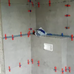 Łazienka w trakcie układania szarych płytek wielkoformatowych, widoczne systemy poziomowania płytek (czerwone i niebieskie klipsy), paczka płytek na zabudowie geberitu, przygotowane otwory...