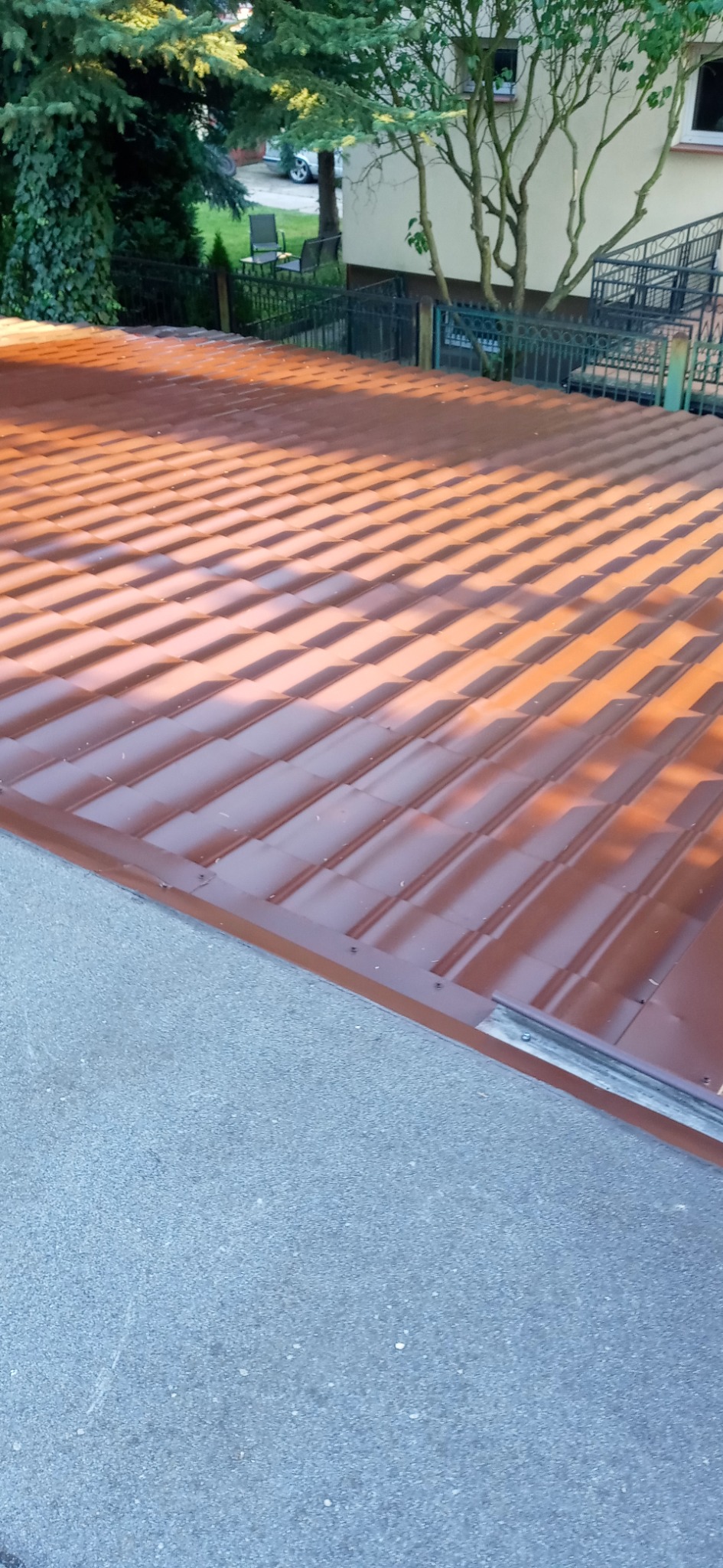 Nowe, brązowe pokrycie dachu z blachy trapezowej, widoczne z góry, z fragmentem chodnika i zieleni w tle.