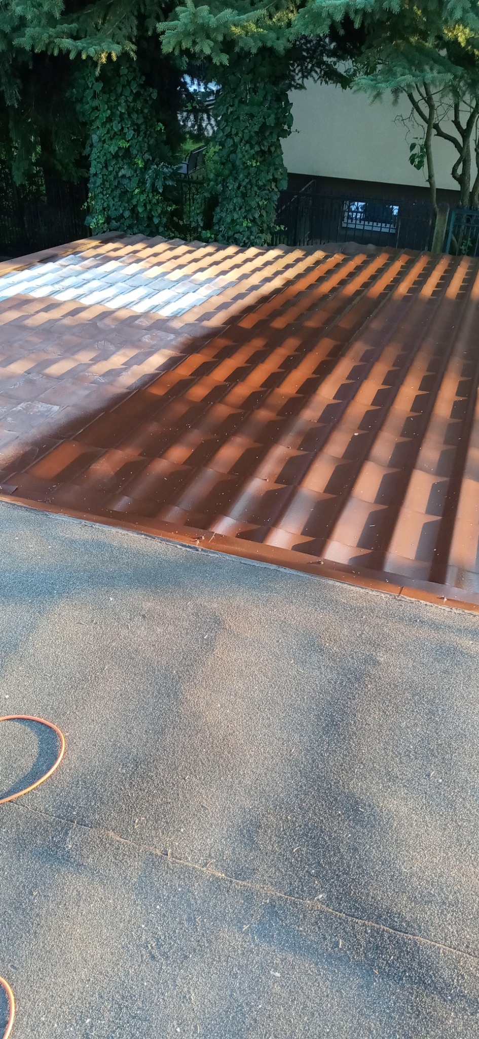 Częściowo pomalowany, brązowy dach pokryty dachówką, z widocznymi śladami świeżej farby i cieniem drzew, sąsiadujący z szarym, płaskim dachem pokrytym papą.