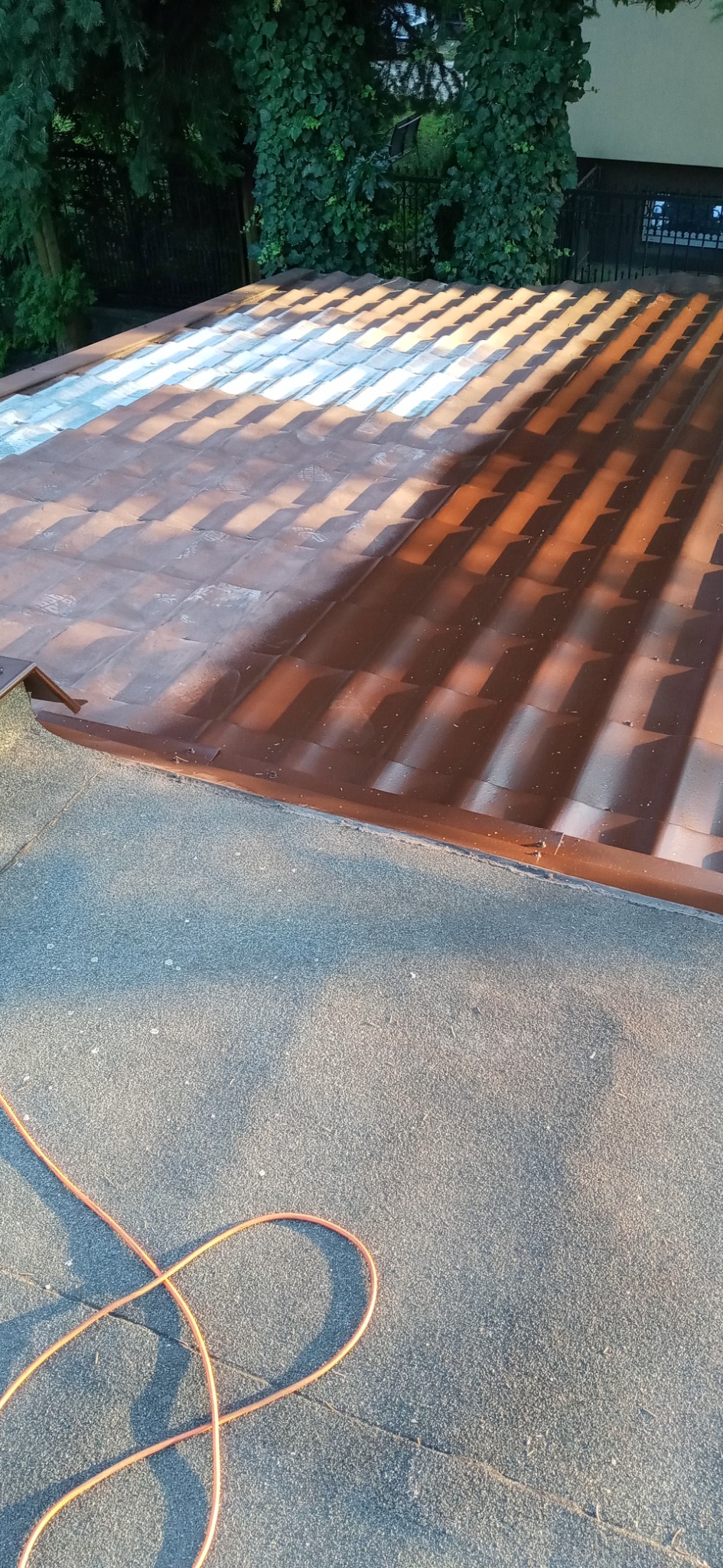 Częściowo pomalowany dach z dachówki ceramicznej, z widocznym fragmentem dachu pokrytego papą i pomarańczowym przewodem na pierwszym planie.