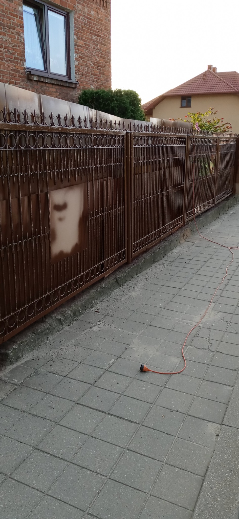 Metalowa brama z dekoracyjnymi elementami, świeżo pomalowana na brązowo, z widocznymi zaciekami farby, na tle ceglanego budynku i kostki brukowej, z pomarańczowym przedłużaczem leżącym na ziemi.