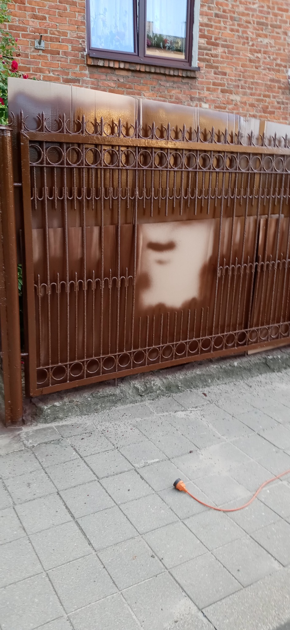 Brama metalowa z dekoracyjnymi elementami, świeżo po nałożeniu brązowej farby, z widocznymi niedomalowaniami i przebarwieniami, na tle ceglanego budynku z oknem i fragmentem czerwonych kwiatów,...