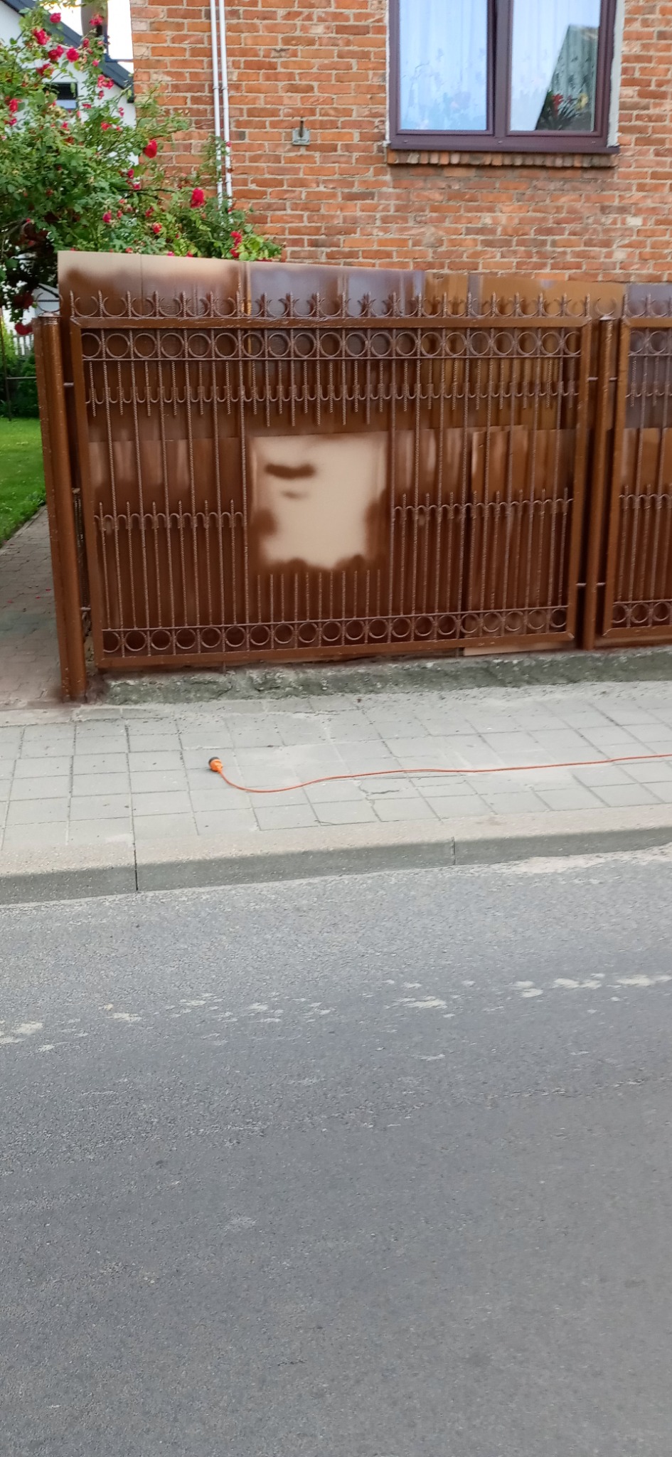Metalowa brama posesji w trakcie renowacji, z widocznymi śladami starej i nowej farby, na tle ceglanego domu z kwitnącymi różami w ogrodzie.