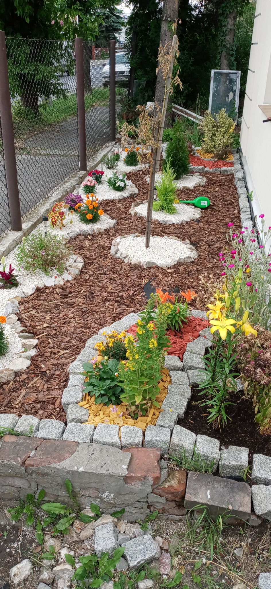 Ogród przydomowy z rabatami kwiatowymi oddzielonymi korą, białym żwirem i obrzeżami z kostki granitowej, widoczna zieleń, kwiaty oraz częściowo zniszczony murek z cegieł.