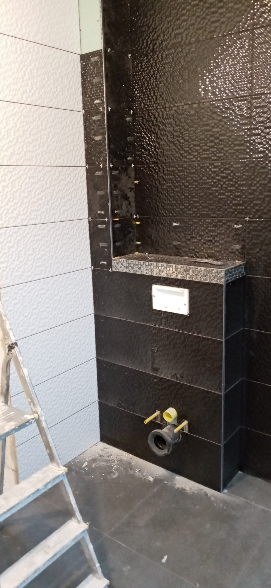 Montaż stelaża podtynkowego WC z czarnymi płytkami o wyraźnej fakturze i białymi płytkami w tle, widoczna drabina malarska, realizacja w Radomiu.