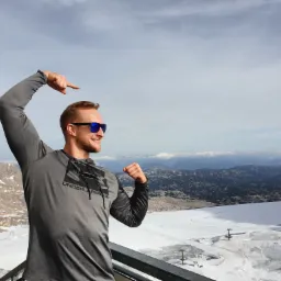 Uśmiechnięty mężczyzna w sportowej koszulce i okularach przeciwsłonecznych, pozujący na tle górskiego krajobrazu pokrytego śniegiem, z uniesioną ręką wskazującą w górę.