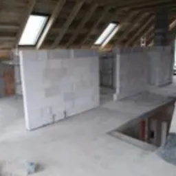 Surowe wnętrze poddasza z oknami dachowymi, ściany z białych bloczków, widoczne drewniane elementy konstrukcji dachu, w trakcie prac budowlanych.