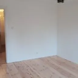 Pusty pokój po remoncie: świeżo pomalowane białe ściany, odnowiona drewniana podłoga z widocznym usłojeniem, wisząca lampa w stylu retro, widok na korytarz przez otwarte drzwi.