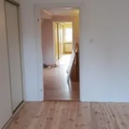 Pusty pokój z drewnianą podłogą w odcieniach beżu i brązu, białe ściany i drzwi prowadzące do kolejnego pomieszczenia z widocznym oknem w tle. Widoczne ślady po niedawno zakończonych pracach.