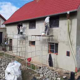 Hurtownia Materiałów i Chemi Budowlanej - HURTBAU - Dwóch pracowników na rusztowaniu podczas prac wykończeniowych elewacji domu jednorodzinnego, zabezpieczone okna folią, widoczne ślady po nakładaniu materiału, na ziemi rozsypana kostka brukowa...