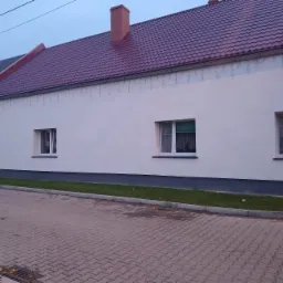 Odnowiona elewacja budynku z nowymi oknami i pomalowanym dachem, widoczne ślady po starej instalacji oświetleniowej.