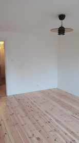 Pusty pokój po remoncie: świeżo pomalowane białe ściany, odnowiona drewniana podłoga z widocznym usłojeniem, wisząca lampa w stylu retro, widok na korytarz przez otwarte drzwi.