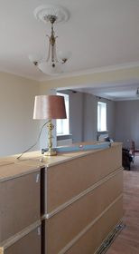 Wnętrze w trakcie remontu: widok na pomieszczenie z drewnianą podłogą, częściowo zasłonięte przez ułożone poziomo płyty kartonowo-gipsowe, na których stoi lampka nocna. W tle widoczne okno...