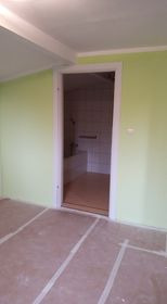 Pusty pokój z zielonymi ścianami w trakcie remontu, widoczne przygotowane podłoże pod posadzkę oraz otwarte drzwi do łazienki z widoczną wanną i płytkami.