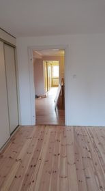 Pusty pokój z drewnianą podłogą w odcieniach beżu i brązu, białe ściany i drzwi prowadzące do kolejnego pomieszczenia z widocznym oknem w tle. Widoczne ślady po niedawno zakończonych pracach.