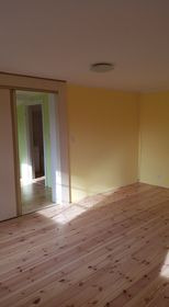 Pusty pokój z drewnianą podłogą i żółtą ścianą, widok na kolejne pomieszczenie przez otwarte przesuwne drzwi, jasne światło słoneczne wpadające przez okno.