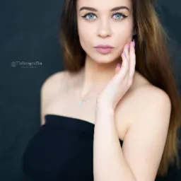 Portret młodej kobiety z długimi brązowymi włosami, błękitnymi oczami i subtelnym makijażem, pozującej na ciemnym tle z dłonią dotykającą policzka, widoczne logo fotografa w lewym dolnym rogu.