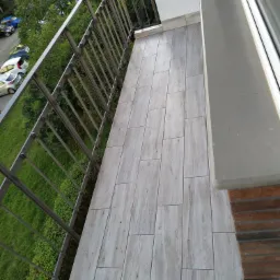 Balkon z płytkami imitującymi drewno, szara metalowa balustrada, widok na zieleń i parking z góry.