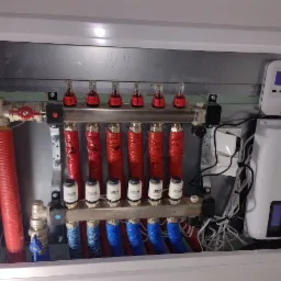 Rozdzielacz ogrzewania podłogowego w szafce, z czerwonymi i niebieskimi rurami, zaworami, przepływomierzami, oraz sterownikiem elektronicznym TECH ST-22.