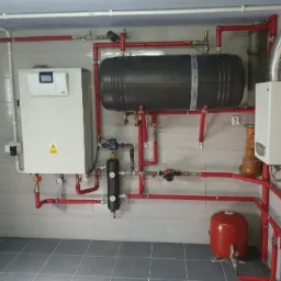 Kotłownia z nowoczesnym piecem, zasobnikiem ciepłej wody i rozbudowaną instalacją z czerwonych rur, widoczne manometry i zawory, szara płytka na ścianie i podłodze.