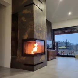 Nowoczesny kominek obłożony płytami imitującymi kamień w salonie z dużym oknem i widokiem na zewnątrz, płomienie widoczne za szybą.