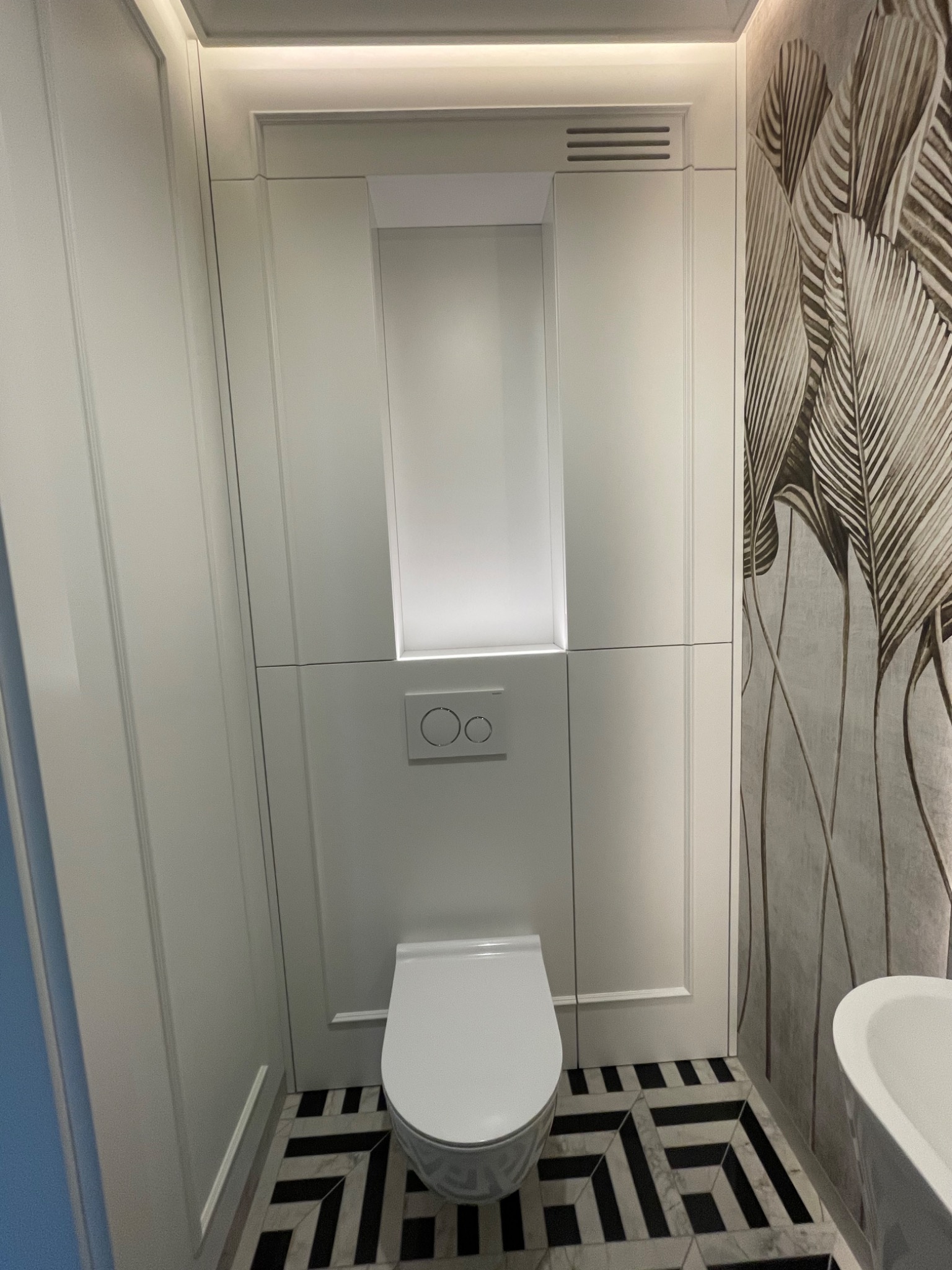 Nowoczesna toaleta podtynkowa z białym panelem przycisków i ukrytą spłuczką, na podłodze wzór w czarno-białą szachownicę, ściana z tapetą w motywie liści.