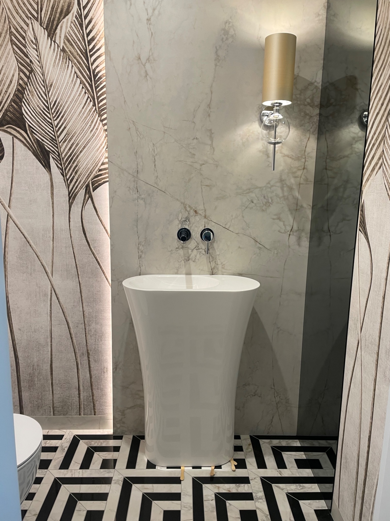 Nowoczesna łazienka z wolnostojącą, białą umywalką na drewnianych nóżkach, marmurową ścianą, wzorzystą tapetą z motywem liści i geometryczną posadzką w czarno-białe wzory.