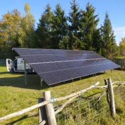 Instalacja paneli słonecznych na metalowej konstrukcji na trawiastym terenie, z otwartym busem serwisowym w tle i drewnianym płotem na pierwszym planie, w słoneczny dzień.