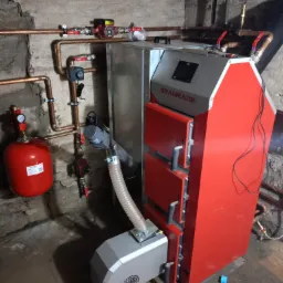 Czerwono-szary piec na paliwo stałe marki Stalmark w kotłowni, z widoczną instalacją hydrauliczną, pompą obiegową i zbiornikiem wyrównawczym.